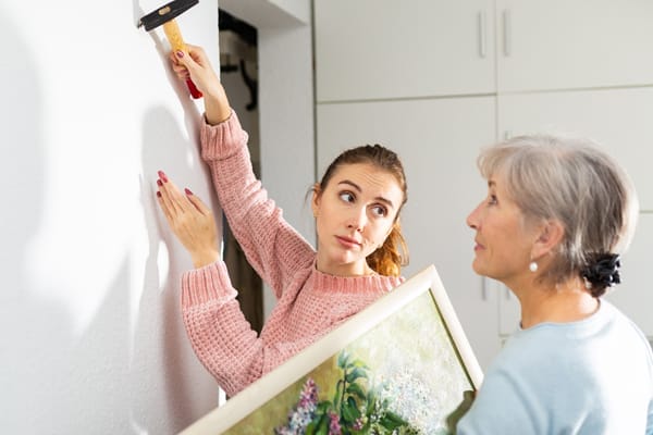 Adult woman helping her senior mother to put painting on wall. Daughter hammering nail, her mother standing beside with picture in hands.
